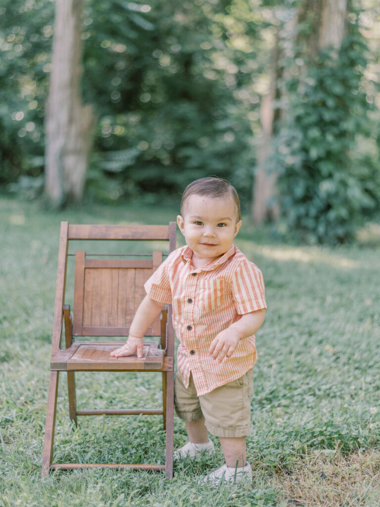 Young boy in button up shirt and white shoes leaning on antique chair at local park.