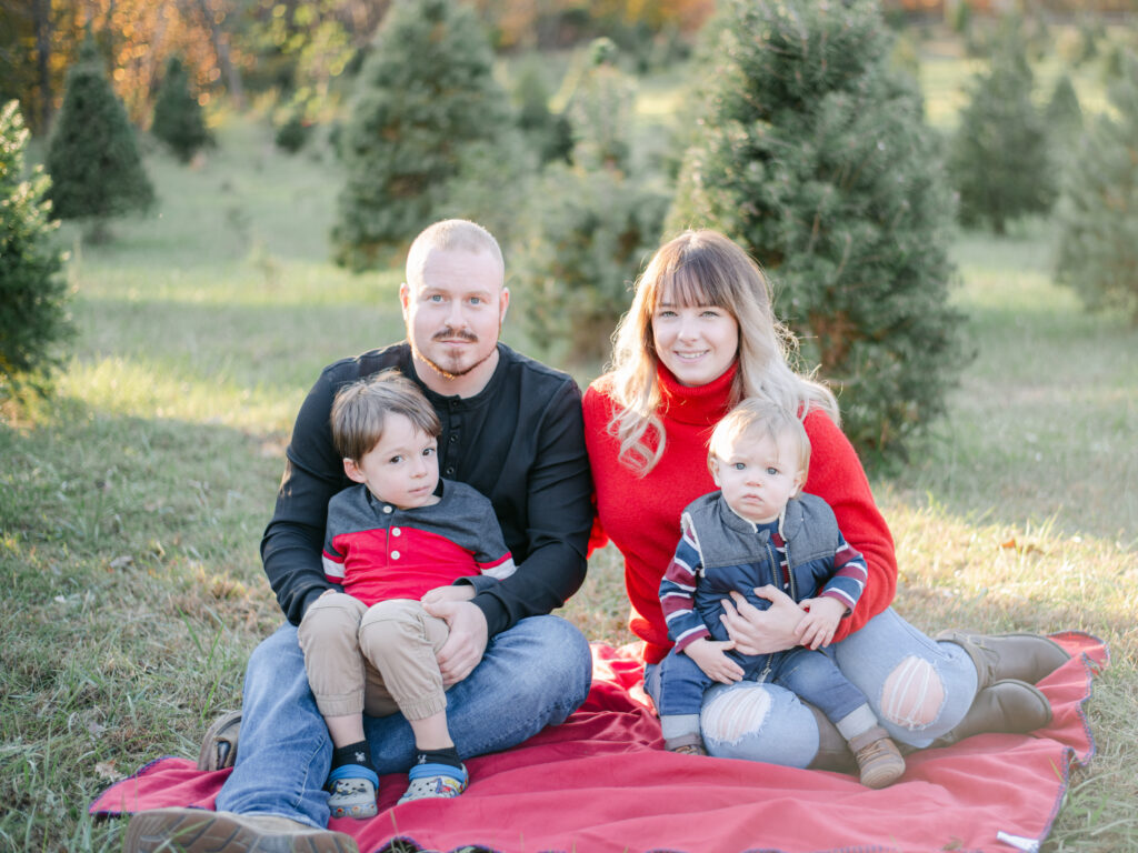 Mom and Dad sitting on red blanket holding sons during Tree Farm photos with Shelby McKimmie Photography