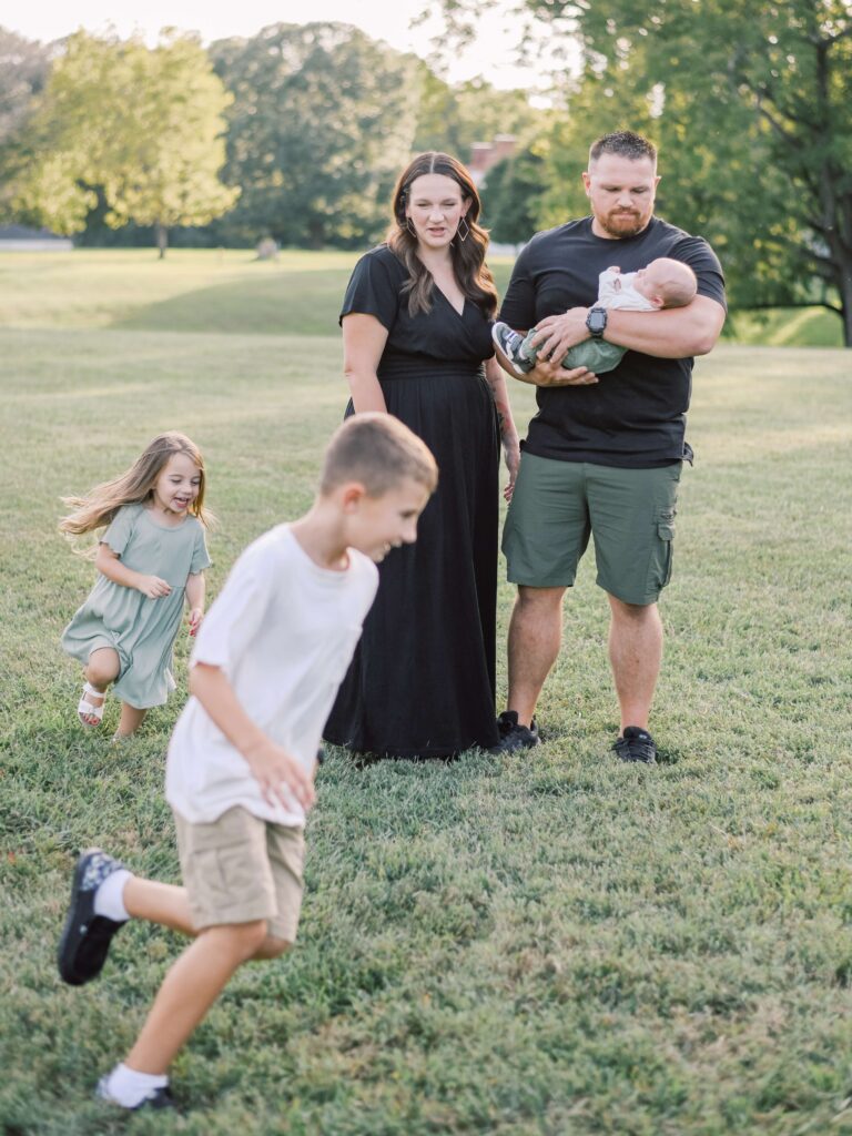 New parents holding their newborn son while older son and daughter play chase.