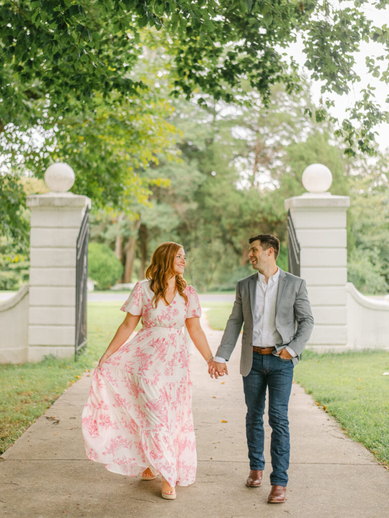 Woman in pink floral dress holding hands with man wearing a gray suit jacket. 
