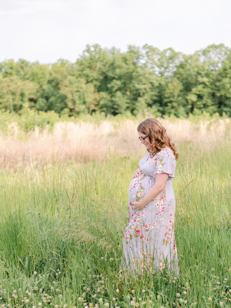 Young mom in floral dress looking at her baby bump in Maryland field.