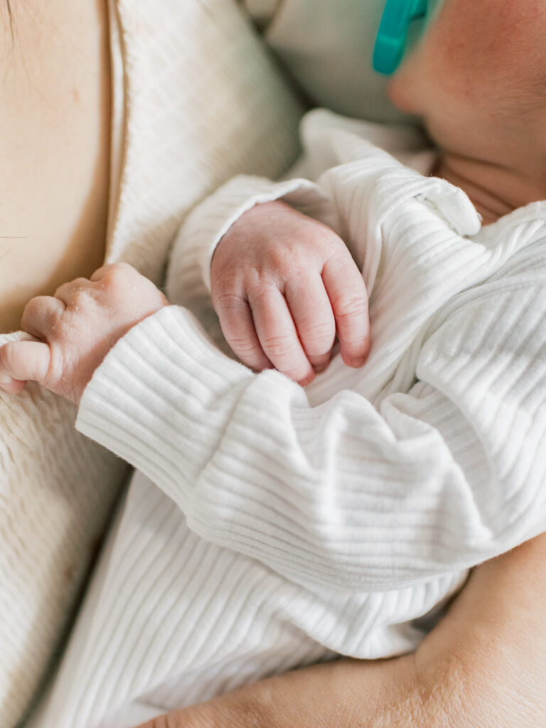 Newborn baby boy wearing a white outfit holding onto his moms dress during a newborn photo session. 