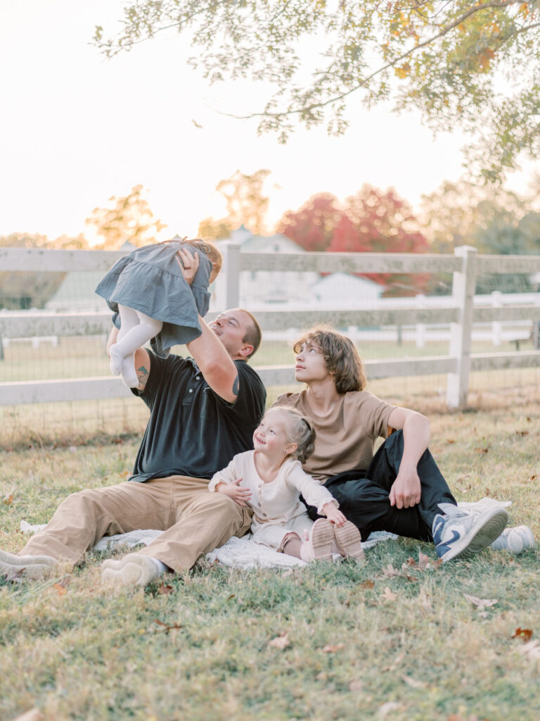 Dad holding his daughter in the air with two other children smiling at daughter.